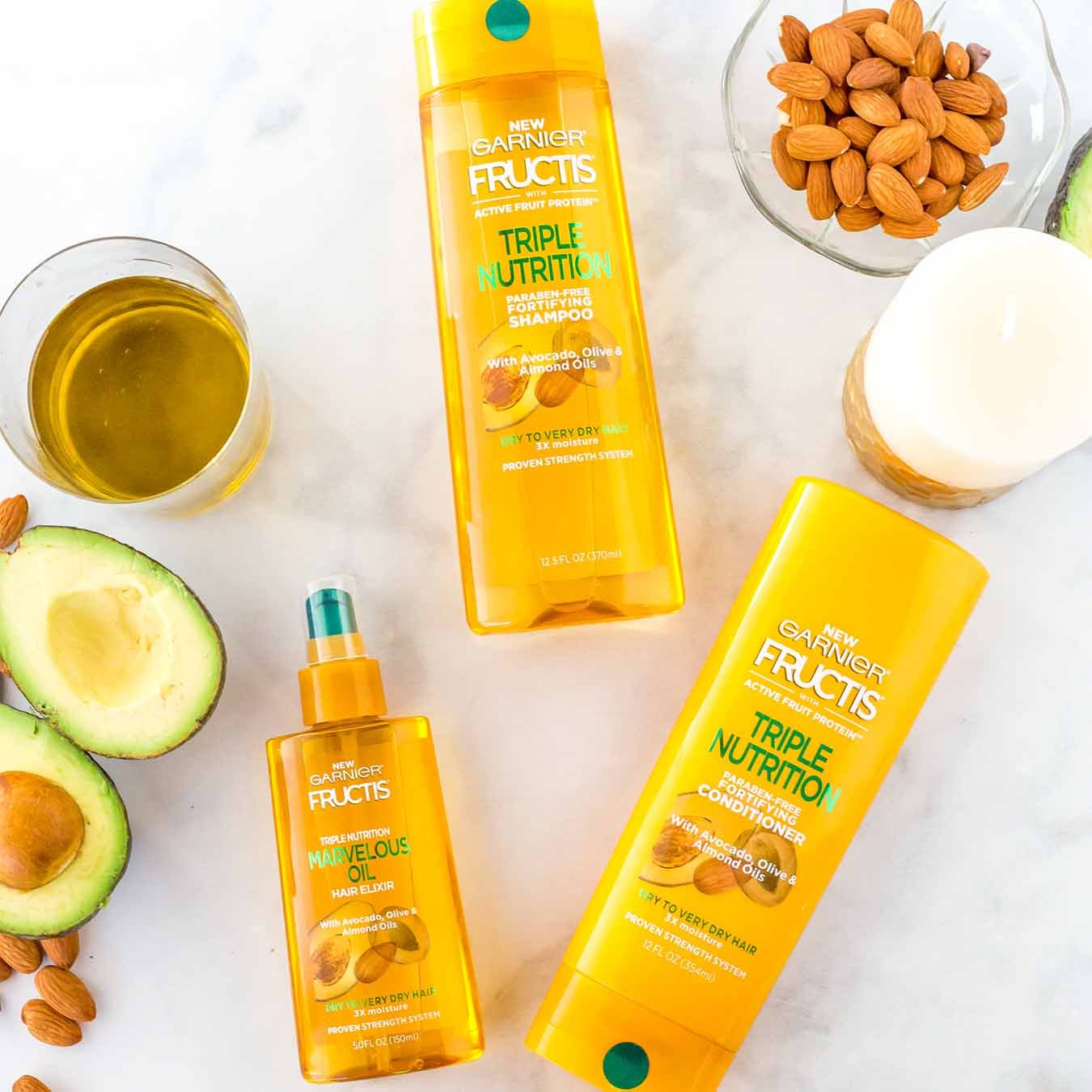 Garnier Fructis Triple Nutrition Shampoo, Fructis Triple Nutrition Conditioner, and Fructis Marvelous Oil on white marble next to halved avocados, a white candle, a glass of oil, a bowl of almonds with some spilled out.